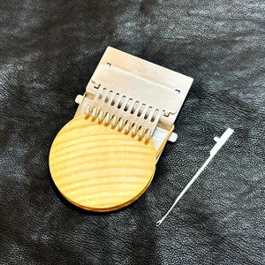 May include: A silver metal loom with a wooden round base and a silver metal hook. The loom is on a black leather surface.