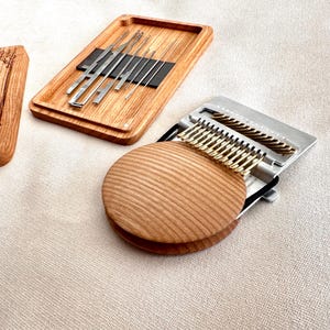 Peut inclure: Un métier à repriser en bois avec un dessus circulaire, une base métallique à crochets et un plateau rectangulaire en bois contenant plusieurs aiguilles à repriser. Une planche en bois gravée DARNING NEEDLES est visible à gauche.