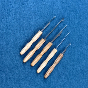 May include: Five crochet hooks with wooden handles and metal tips are arranged diagonally on a blue surface. The handles vary in colour from light to dark wood tones. The hooks are designed for crafting and textile work.
