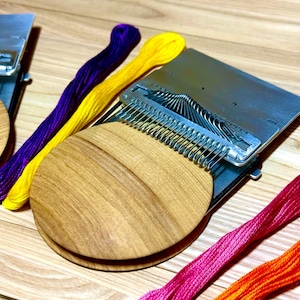 Op de afbeelding: Een ambachtelijke weefgereedschapsset met houten bases en metalen componenten. Kleurrijk borduurgaren in paars, geel, roze en oranje is rond de gereedschappen gerangschikt. De houten bases zijn ovaal van vorm.