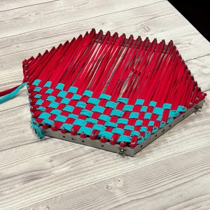 May include: A woven craft project with a geometric design. The piece features a metal frame and is filled with red and turquoise fabric strips. The strips are woven together in a basket-weave pattern, creating a textured surface.