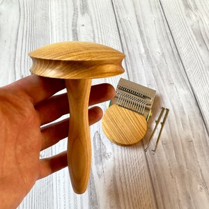 Puede incluir: Un telar de punto de madera con un mango en forma de hongo y una base circular. El telar est&aacute; hecho de madera marr&oacute;n claro y tiene una aguja de metal plateado.