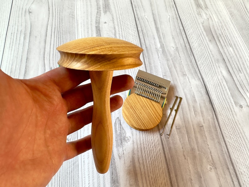 Puede incluir: Un telar de punto de madera con un mango en forma de hongo y una base circular. El telar est&aacute; hecho de madera marr&oacute;n claro y tiene una aguja de metal plateado.