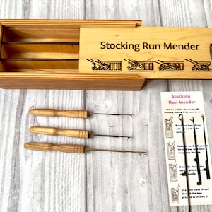 Op de afbeelding: Een houten doos met een deksel dat opengaat om drie breinaalden met houten handvatten te onthullen. De doos is gelabeld "Stocking Run Mender" en heeft een diagram van hoe de naalden te gebruiken. Een aparte kaart met instructies is ook inbegrepen.
