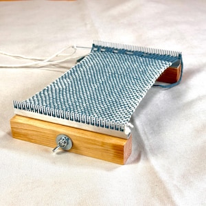 Peut inclure: Un métier à tisser en bois composé de deux blocs de bois clair reliés par des tiges métalliques. Un morceau de tissu partiellement tissé avec un motif à carreaux bleu et blanc est tendu entre les deux peignes métalliques. Du fil blanc dépasse.