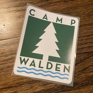May include: A green and white sticker with the text "CAMP WALDEN" and a white pine tree graphic. The text "CAMP" is in all caps and the text "WALDEN" is in all caps. There are three blue wavy lines at the bottom of the sticker.