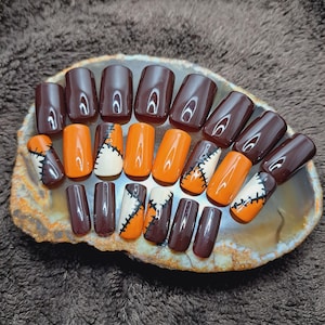 May include: Set of 14 false nails with a Halloween theme. The nails are brown, orange, and white with a stitched design.