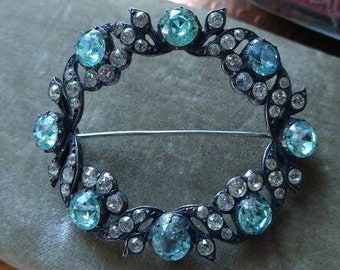 May include: A silver and black brooch with a circular design. The brooch is set with a row of light blue and clear rhinestones.