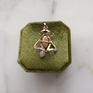 May include: A gold-toned metal pendant featuring an openwork frame with a central oval cabochon and a small, dangling, pale, iridescent drop-shaped bead at the bottom. The piece includes a top loop for attachment to a chain or cord.