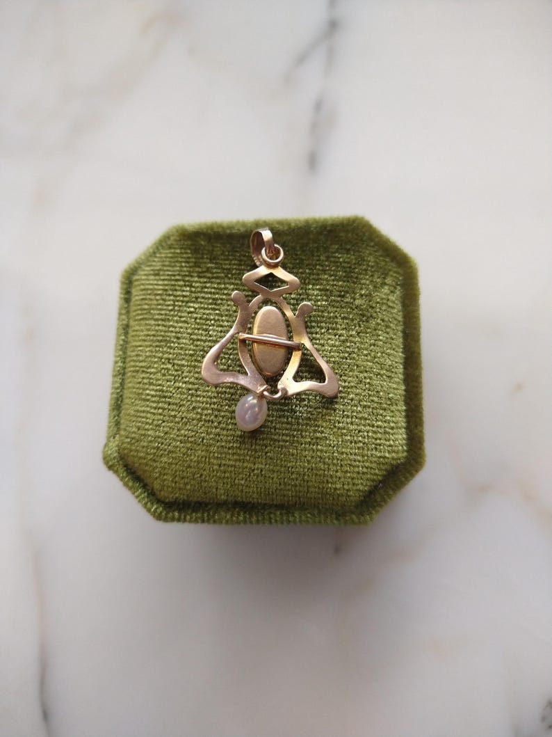 May include: A gold-toned metal pendant featuring an openwork frame with a central oval cabochon and a small, dangling, pale, iridescent drop-shaped bead at the bottom. The piece includes a top loop for attachment to a chain or cord.