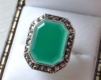 May include: A silver ring with a large, emerald-green, octagonal gemstone. The gemstone is surrounded by a border of small, dark, sparkling stones. The ring is set in a white box.