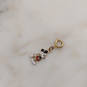 May include: A gold charm depicting Mickey Mouse, with black, red, white, and yellow enamel detailing. It has a clasp for attaching to a bracelet or necklace. The charm is from The Old Jewelry Chest. The charm is gold in colour.