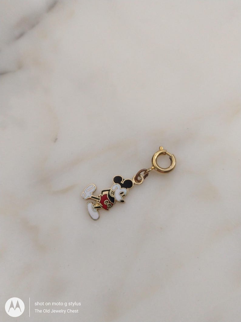 May include: A gold charm depicting Mickey Mouse, with black, red, white, and yellow enamel detailing. It has a clasp for attaching to a bracelet or necklace. The charm is from The Old Jewelry Chest. The charm is gold in colour.
