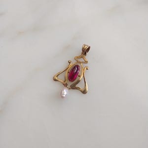 May include: A gold-toned pendant featuring a central oval-shaped deep red gemstone set within a curved, openwork metal frame. A small, irregular white pearl hangs from the bottom of the metal structure, which includes a top loop for a chain.