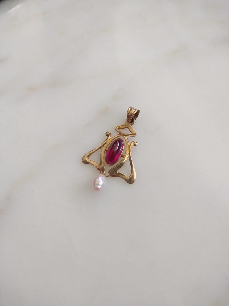May include: A gold-toned pendant featuring a central oval-shaped deep red gemstone set within a curved, openwork metal frame. A small, irregular white pearl hangs from the bottom of the metal structure, which includes a top loop for a chain.
