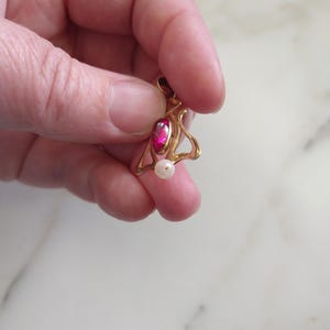 May include: A gold pendant featuring a teardrop-shaped deep pink gemstone set in a curved metal frame with a small white spherical pearl dangling from the bottom. The pendant has a small circular bail at the top for attachment to a chain.