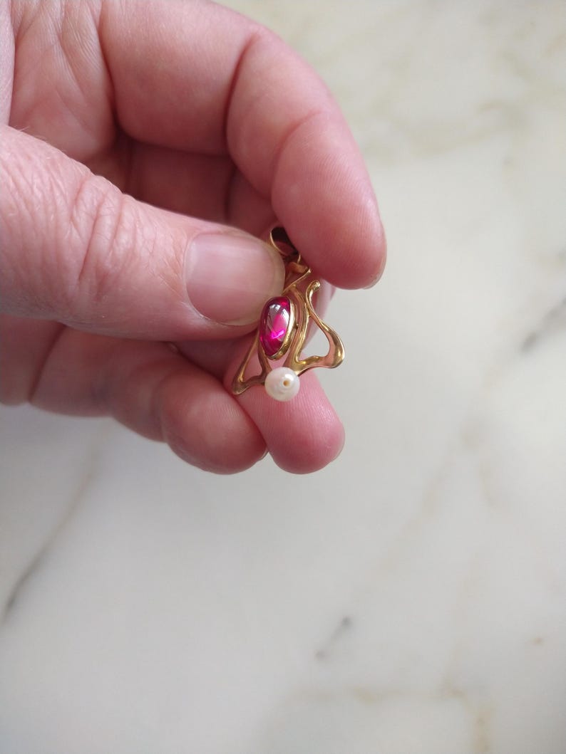 May include: A gold pendant featuring a teardrop-shaped deep pink gemstone set in a curved metal frame with a small white spherical pearl dangling from the bottom. The pendant has a small circular bail at the top for attachment to a chain.