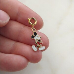 May include: A gold-coloured Mickey Mouse charm with black ears, a white face, and a red outfit. The charm is attached to a gold-coloured clasp. The charm is a small, detailed piece of jewellery.
