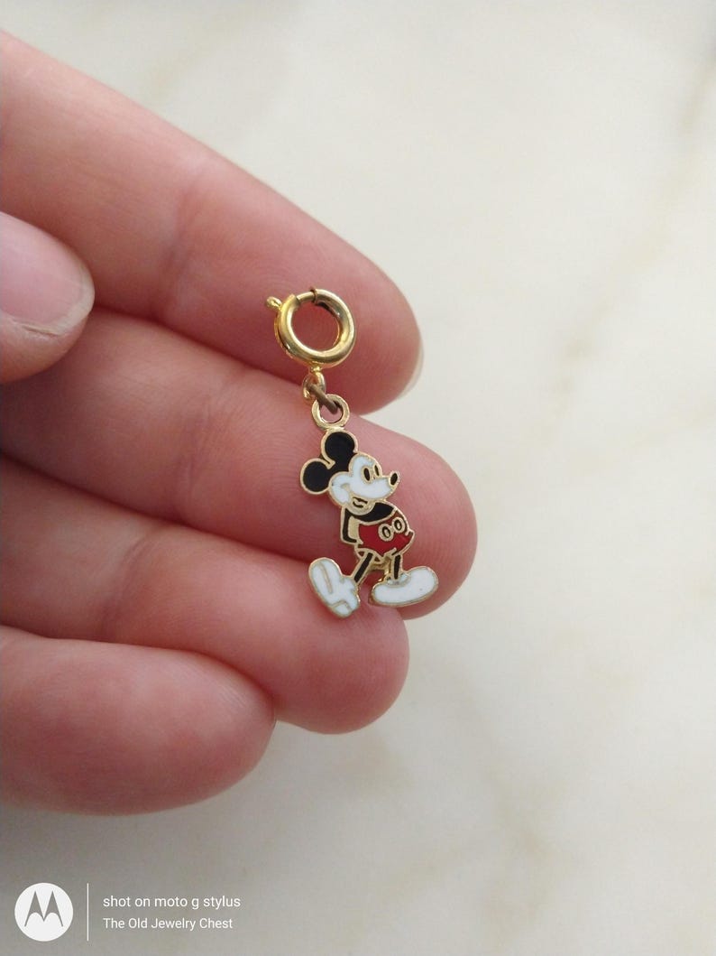 May include: A gold-coloured Mickey Mouse charm with black ears, a white face, and a red outfit. The charm is attached to a gold-coloured clasp. The charm is a small, detailed piece of jewellery.