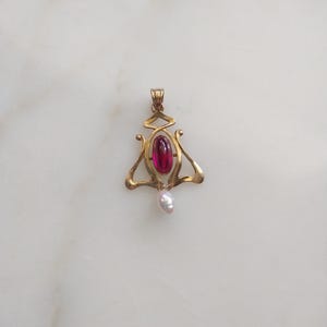 May include: A gold pendant featuring a central oval-shaped deep pink gemstone and a small white teardrop-shaped pearl dangling from the bottom. The metal frame has a curved, openwork design with a bail at the top for attaching to a chain.