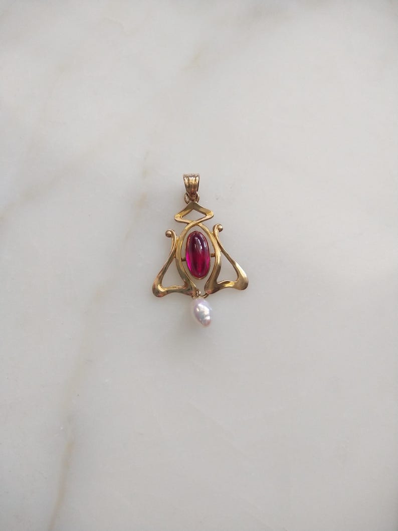 May include: A gold pendant featuring a central oval-shaped deep pink gemstone and a small white teardrop-shaped pearl dangling from the bottom. The metal frame has a curved, openwork design with a bail at the top for attaching to a chain.