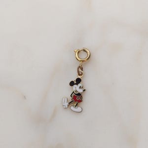 May include: A gold-coloured charm with a colourful enamel Mickey Mouse design. The charm has a red shirt, white gloves and shoes, and black ears. The charm is attached to a gold-coloured clasp.