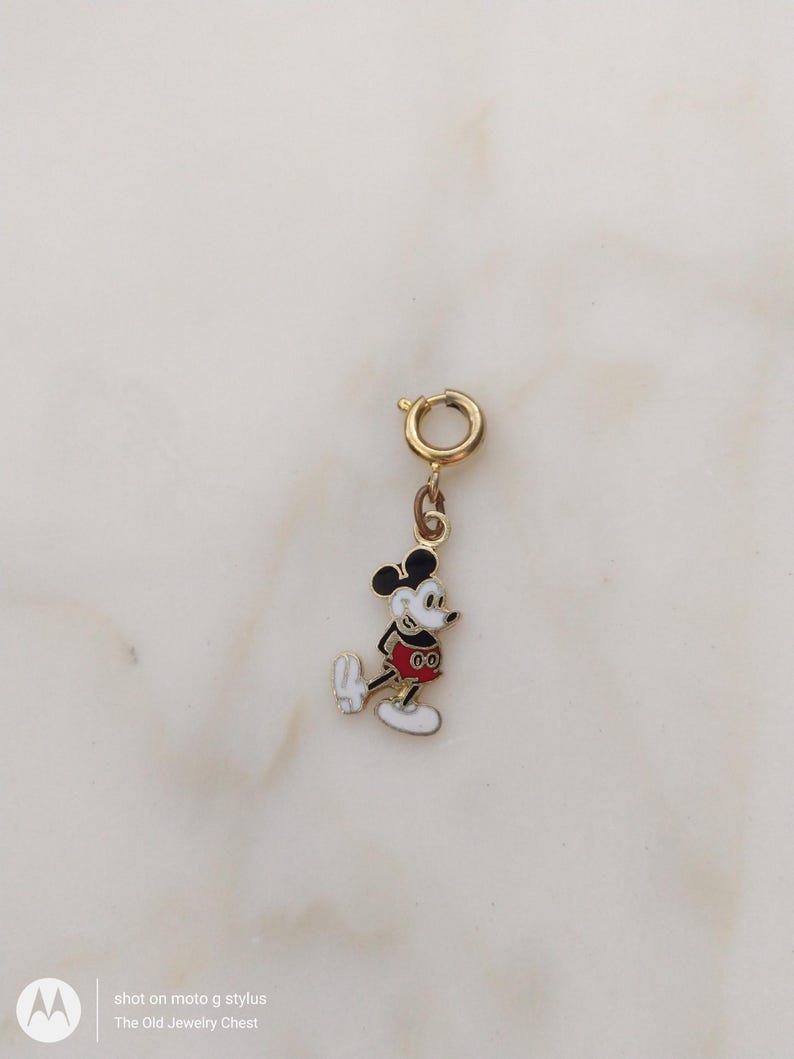 May include: A gold-coloured charm with a colourful enamel Mickey Mouse design. The charm has a red shirt, white gloves and shoes, and black ears. The charm is attached to a gold-coloured clasp.