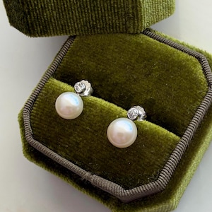 May include: A pair of stud earrings featuring round white pearls set in silver-toned metal, each accented with a single small clear round-cut gemstone positioned directly above the pearl. The earrings are displayed inside a green velvet jewelry box.