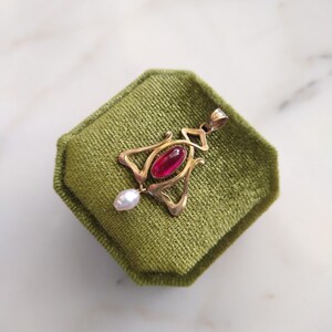 May include: A gold pendant featuring a central oval-shaped deep red gemstone and a small white teardrop-shaped pearl dangling from the bottom. The metal frame has an openwork design with curved lines and a bail at the top for a necklace chain.