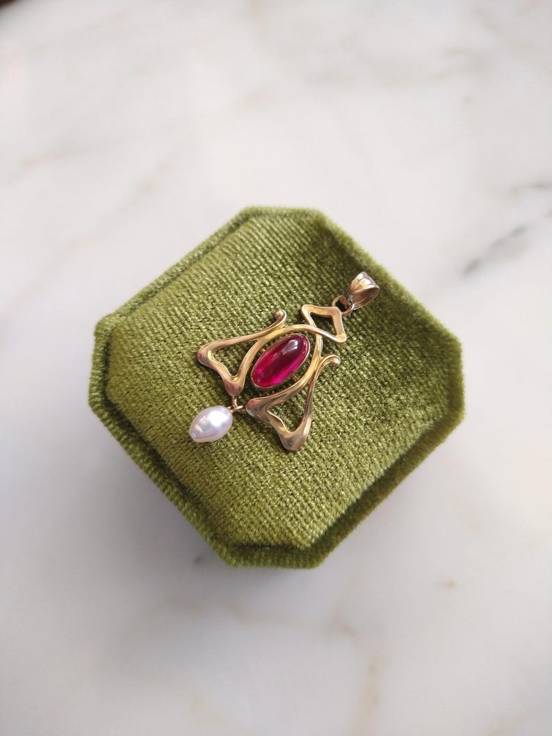 May include: A gold pendant featuring a central oval-shaped deep red gemstone and a small white teardrop-shaped pearl dangling from the bottom. The metal frame has an openwork design with curved lines and a bail at the top for a necklace chain.