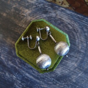 May include: A pair of silver-toned clip-on earrings with textured, spherical drops. The earrings are displayed on a square, green velvet surface. The earrings have a screw-back mechanism for secure wear.