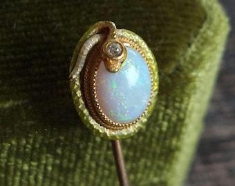 May include: A gold stick pin featuring an oval-shaped opal cabochon set within a textured gold border. A small gold snake with a tiny diamond set in its head wraps around the top edge of the opal, which displays iridescent flashes of color.