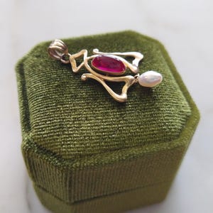 May include: A gold pendant featuring a central oval-shaped deep pink gemstone and a small white teardrop-shaped pearl dangling from the bottom. The metal frame has an openwork design with a bail at the top for attaching to a necklace chain.