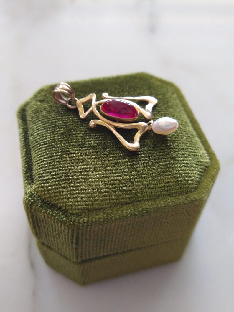 May include: A gold pendant featuring a central oval-shaped deep pink gemstone and a small white teardrop-shaped pearl dangling from the bottom. The metal frame has an openwork design with a bail at the top for attaching to a necklace chain.