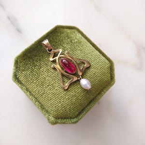 May include: A gold pendant featuring an elongated, oval-shaped deep pink gemstone set in the center of a stylized, open-work metal frame. A small, teardrop-shaped white pearl hangs from the bottom of the gold structure, which includes a bail.