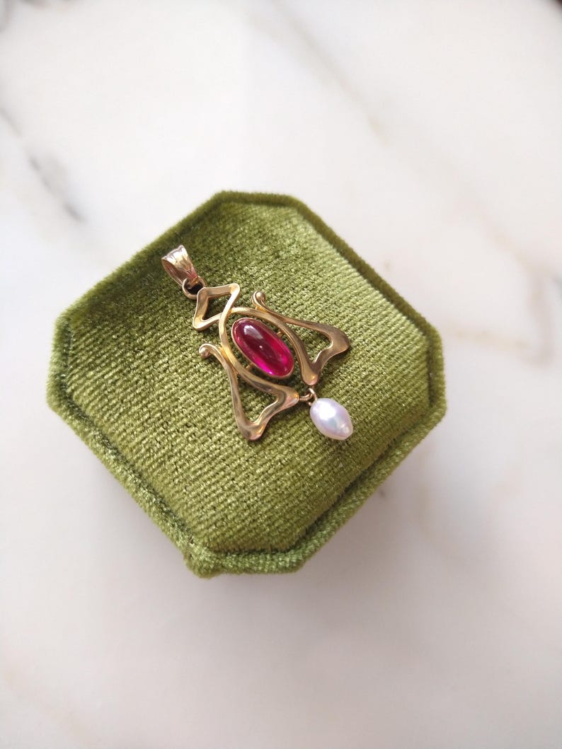May include: A gold pendant featuring an elongated, oval-shaped deep pink gemstone set in the center of a stylized, open-work metal frame. A small, teardrop-shaped white pearl hangs from the bottom of the gold structure, which includes a bail.
