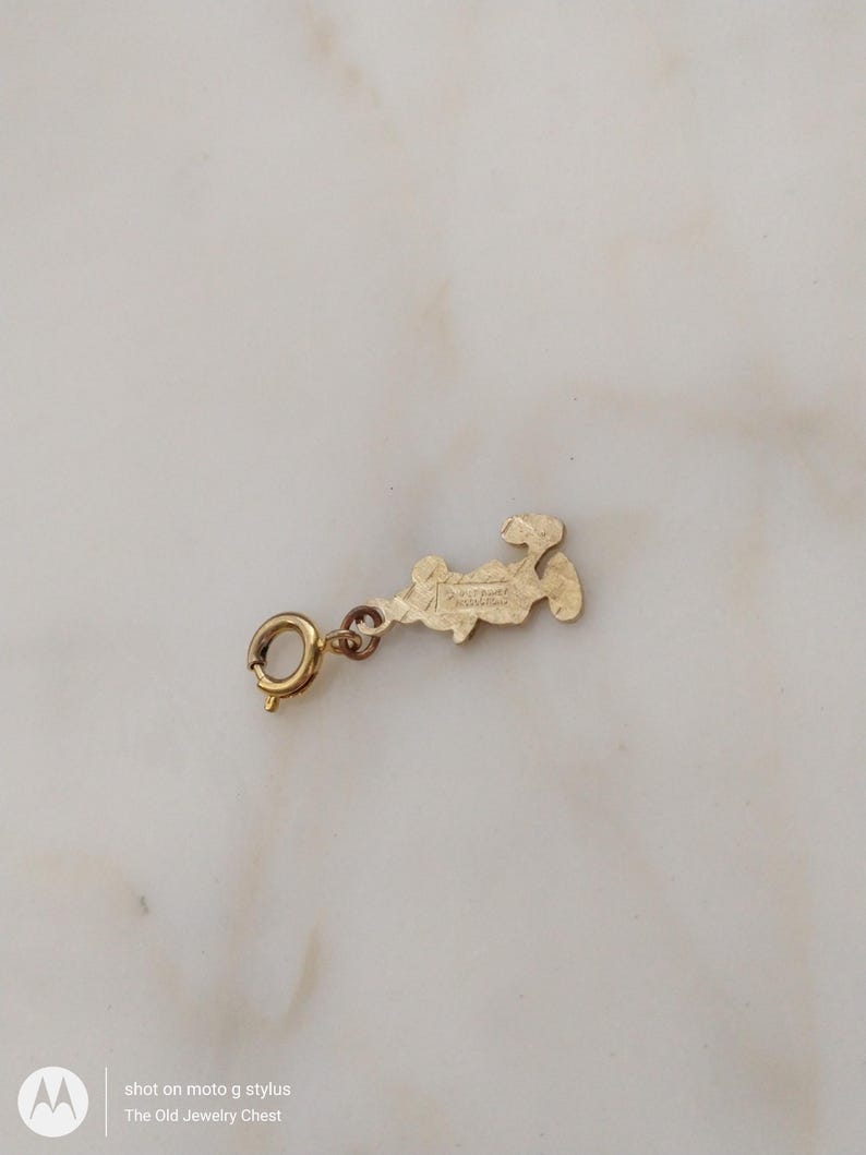 May include: A gold-coloured charm with a small clasp. The charm is in the shape of a stylized figure. The text "The Old Jewelry Chest" is visible in the image.