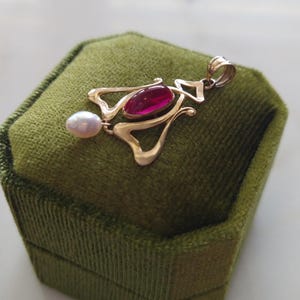 May include: A gold pendant featuring a central oval-shaped deep pink gemstone and a small white teardrop-shaped pearl dangling from the bottom. The metal frame has an openwork design with a polished finish and a top loop for a chain attachment.