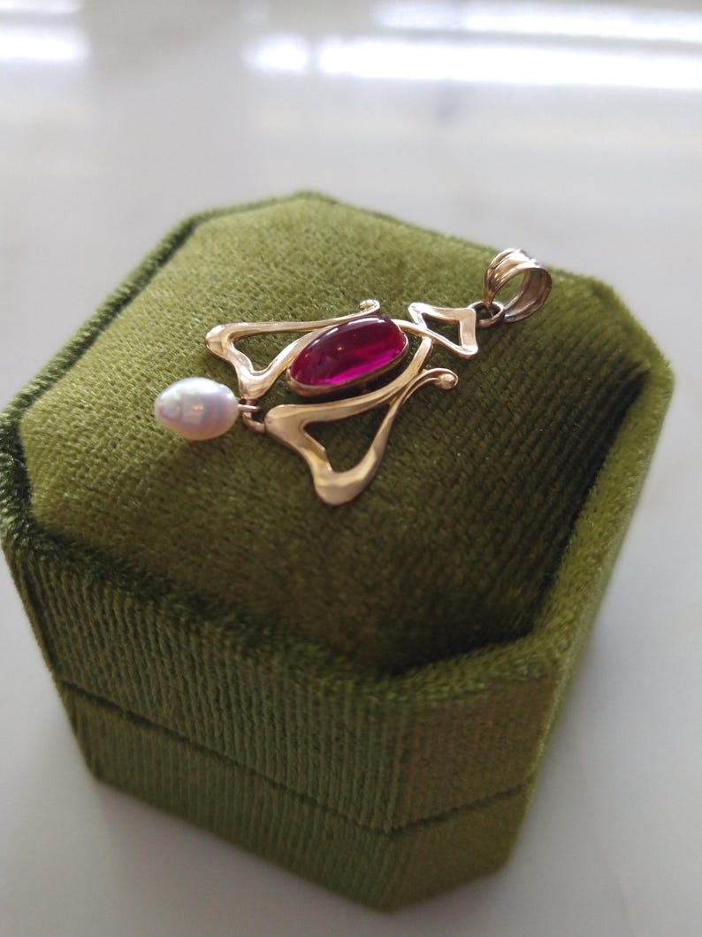May include: A gold pendant featuring a central oval-shaped deep pink gemstone and a small white teardrop-shaped pearl dangling from the bottom. The metal frame has an openwork design with a polished finish and a top loop for a chain attachment.