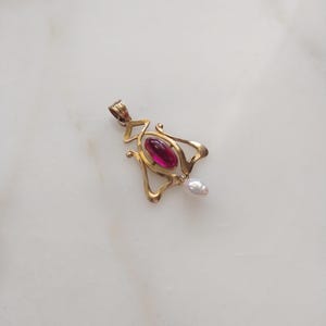 May include: A gold pendant featuring a central oval-shaped deep pink cabochon stone set within a curved metal frame, with a small white teardrop-shaped pearl dangling from the bottom and a metal loop at the top for attachment to a chain.