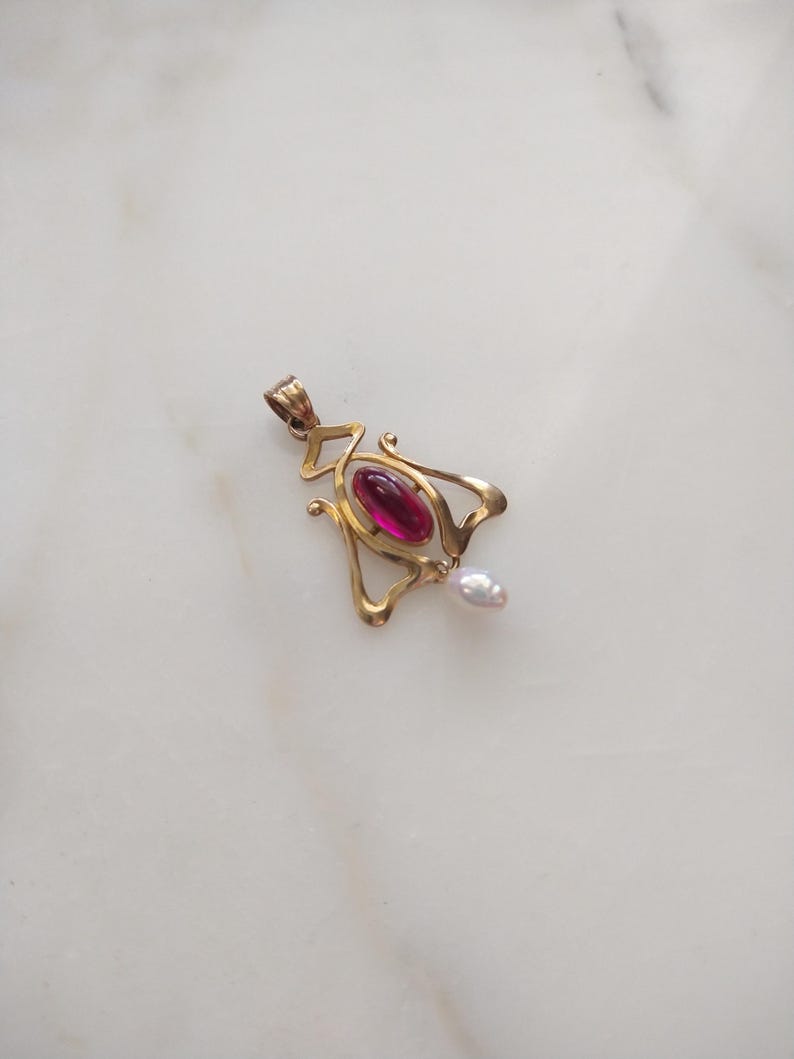 May include: A gold pendant featuring a central oval-shaped deep pink cabochon stone set within a curved metal frame, with a small white teardrop-shaped pearl dangling from the bottom and a metal loop at the top for attachment to a chain.