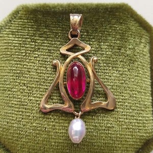 May include: A gold pendant featuring a central oval-shaped cabochon red gemstone set within a polished gold frame with curved, openwork metal detailing. A single white teardrop-shaped pearl hangs from the bottom of the gold metal structure.