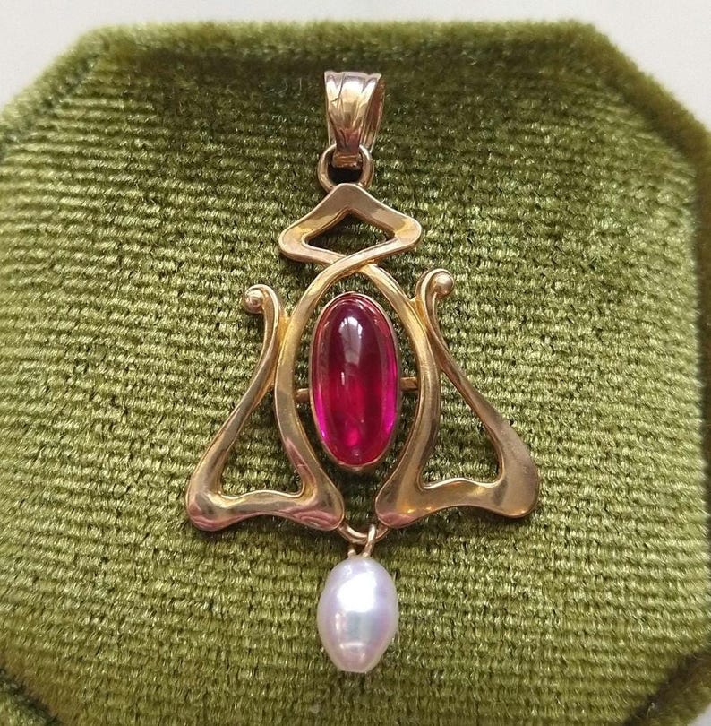 May include: A gold pendant featuring a central oval-shaped cabochon red gemstone set within a polished gold frame with curved, openwork metal detailing. A single white teardrop-shaped pearl hangs from the bottom of the gold metal structure.