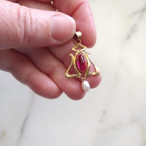 May include: A gold pendant featuring a central oval-shaped deep pink gemstone set within a curved, openwork metal frame. A small, white, teardrop-shaped pearl hangs from the bottom of the gold structure, which includes a top bail for attachment.