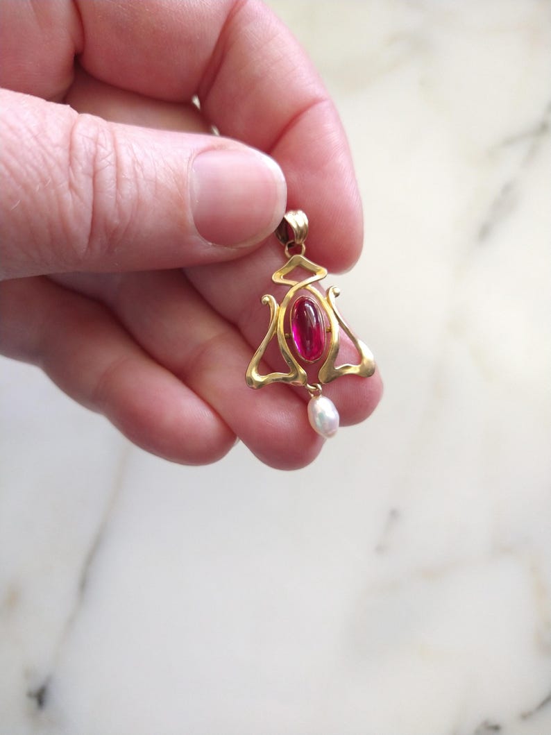 May include: A gold pendant featuring a central oval-shaped deep pink gemstone set within a curved, openwork metal frame. A small, white, teardrop-shaped pearl hangs from the bottom of the gold structure, which includes a top bail for attachment.