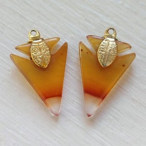 May include: A pair of arrowhead shaped earrings with gold accents. The earrings are made of agate and have a translucent orange and white color.