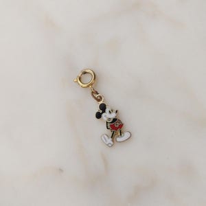 May include: A gold-coloured Mickey Mouse charm with black ears, a red shirt, and white gloves and shoes. The charm has a small clasp for attaching to a bracelet or necklace. The charm is a classic Disney character.
