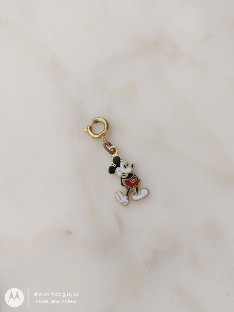 May include: A gold-coloured Mickey Mouse charm with black ears, a red shirt, and white gloves and shoes. The charm has a small clasp for attaching to a bracelet or necklace. The charm is a classic Disney character.