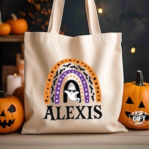 May include: A white canvas tote bag with a rainbow design featuring orange, purple and white stripes, bats and stars. A ghost with sunglasses is in the centre of the rainbow. The text "ALEXIS" is printed in black below the rainbow.