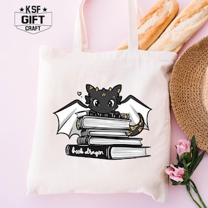 Pu&ograve; includere: Una borsa tote bianca con manici neri presenta un drago di cartone animato nero e oro seduto su una pila di libri. Il testo "book dragon" &egrave; stampato sotto il drago. La borsa ha un piccolo logo bianco e nero che dice "KSF GIFT CRAFT" nell'angolo in alto a sinistra.